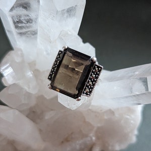 May include: A silver ring with a rectangular, dark brown gemstone set in a silver frame. The ring is surrounded by small, black gemstones.