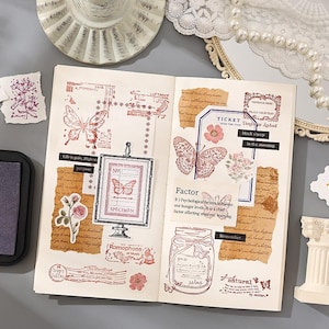 May include: Open journal pages with vintage-style stamps and stickers. The stamps feature butterflies, flowers, and text in red ink. The journal includes a framed butterfly specimen and a jar with a butterfly design.