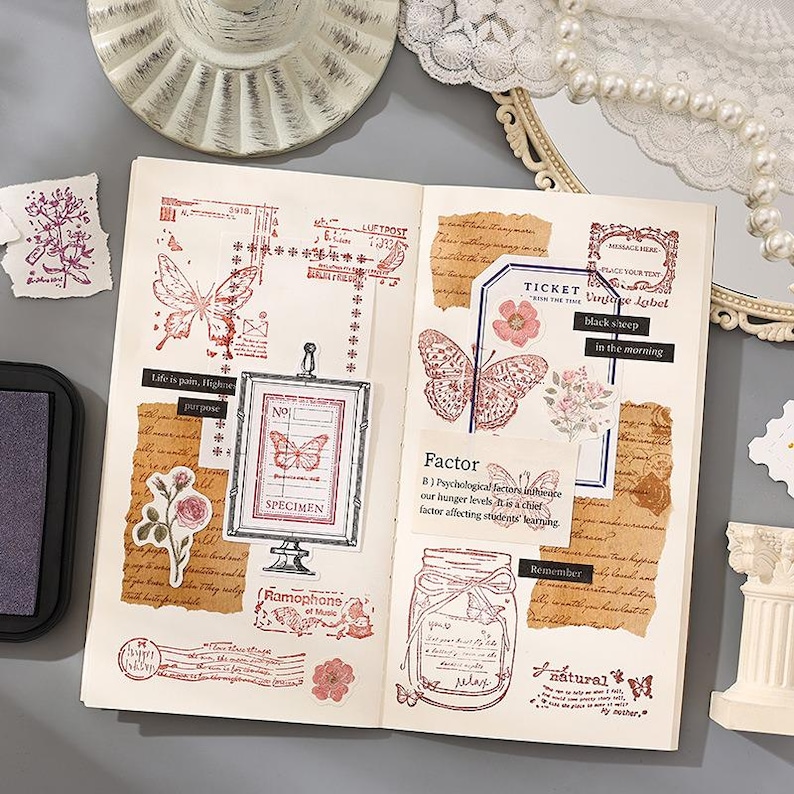 May include: Open journal pages with vintage-style stamps and stickers. The stamps feature butterflies, flowers, and text in red ink. The journal includes a framed butterfly specimen and a jar with a butterfly design.