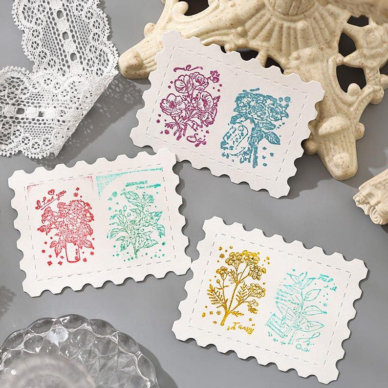 May include: Four white, postage stamp-shaped cards with floral designs in various colors. The cards feature images of flowers and foliage in shades of red, purple, blue, green, and gold. Some cards have text, including "I love you" and other script.