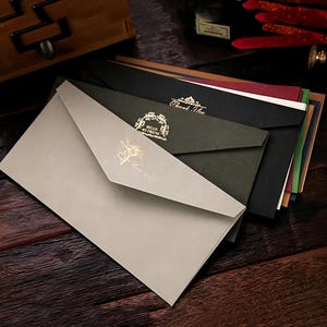 Könnte beinhalten: Ein Stapel eleganter Umschläge in verschiedenen Farben, darunter Grau, Schwarz und Grün, mit goldfarbenen Akzenten. Einige Umschläge tragen die Aufschriften "Hello My Friend" und "Thank You". Die Umschläge liegen auf einer dunklen Holzoberfläche.