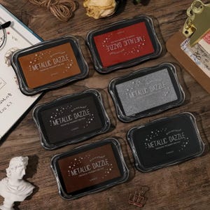 May include: Six rectangular ink pads labelled Metallic Dazzle Premium Pigment Ink Pad in various colours including copper, red, silver, black, and dark brown. Each ink pad is encased in a clear plastic container with a wavy edge design.
