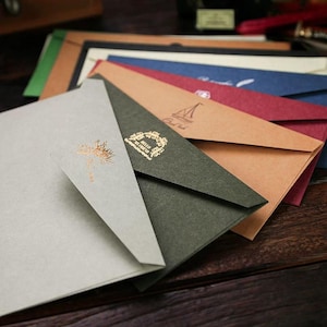 Op de afbeelding: Een verzameling kleurrijke enveloppen in verschillende tinten, waaronder grijs, groen, bruin, rood en blauw. Sommige enveloppen hebben gouden folieontwerpen, zoals een boom en de woorden "Hello My Friend". De enveloppen zijn gerangschikt op een houten oppervlak.