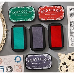 May include: A collection of ink pads in cyan, red, and gray, each labeled with its color name. The pads are rectangular with rounded corners and black bases. The text "color space" is also visible on the labels. The image also includes decorative elements.