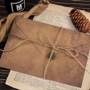 May include: A vintage-style brown envelope tied with twine and a metal four-leaf clover charm. The envelope has a distressed, aged appearance, with a dark ink bottle and dried leaves in the background. The background includes a vintage paper with text.