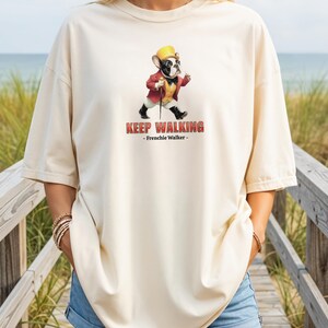 May include: Cream-colored t-shirt featuring a cartoon French Bulldog dressed in a red jacket, yellow vest, and hat, holding a cane. The text "KEEP WALKING - Frenchie Walker-" is printed in red.