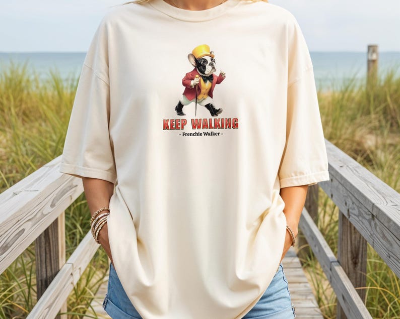 May include: Cream-colored t-shirt featuring a cartoon French Bulldog dressed in a red jacket, yellow vest, and hat, holding a cane. The text "KEEP WALKING - Frenchie Walker-" is printed in red.