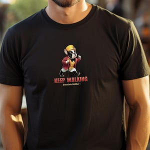May include: Black t-shirt featuring a cartoon French Bulldog wearing a yellow hat and red jacket, walking with a cane. The text "KEEP WALKING - Frenchie Walker -" is printed below the graphic. The shirt is made of a soft material.