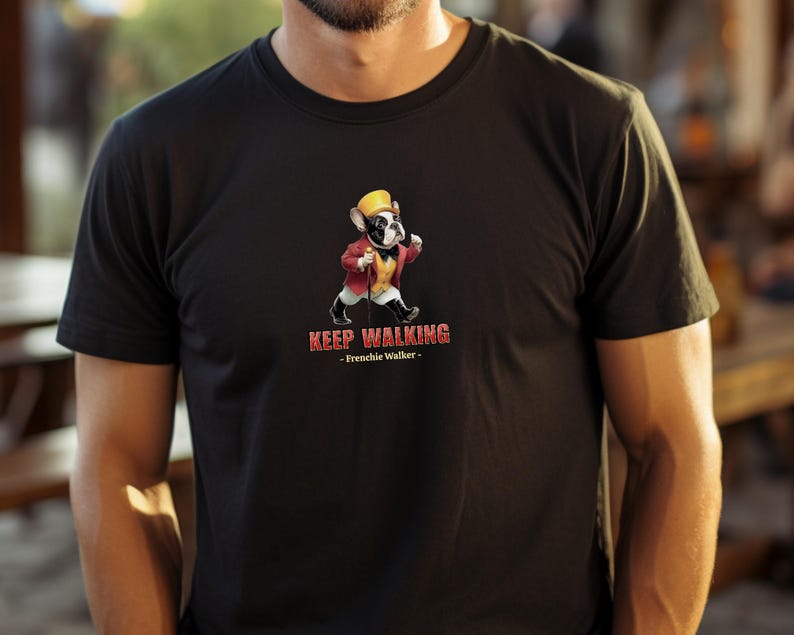 May include: Black t-shirt featuring a cartoon French Bulldog wearing a yellow hat and red jacket, walking with a cane. The text "KEEP WALKING - Frenchie Walker -" is printed below the graphic. The shirt is made of a soft material.