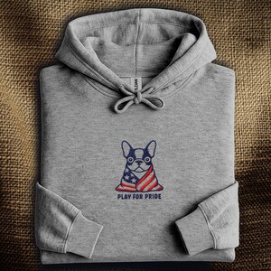 以下が含まれることがあります： アメリカ国旗をまとったフレンチブルドッグの刺繍が施された、ヘザーグレーのパーカー。犬の下には「PLAY FOR PRIDE」の文字。パーカーには、ドローストリングフードとリブ編みの袖口が付いています。
