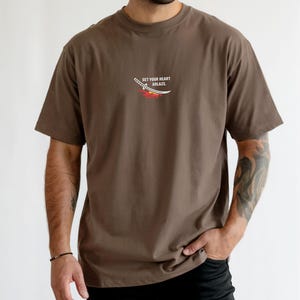 Puede incluir: Una camiseta marr&oacute;n de manga corta con un peque&ntilde;o gr&aacute;fico en el pecho. El gr&aacute;fico muestra una espada katana blanca sobre una llama estilizada roja y naranja, con el texto "SET YOUR HEART ABLAZE" impreso en letras may&uacute;sculas blancas.