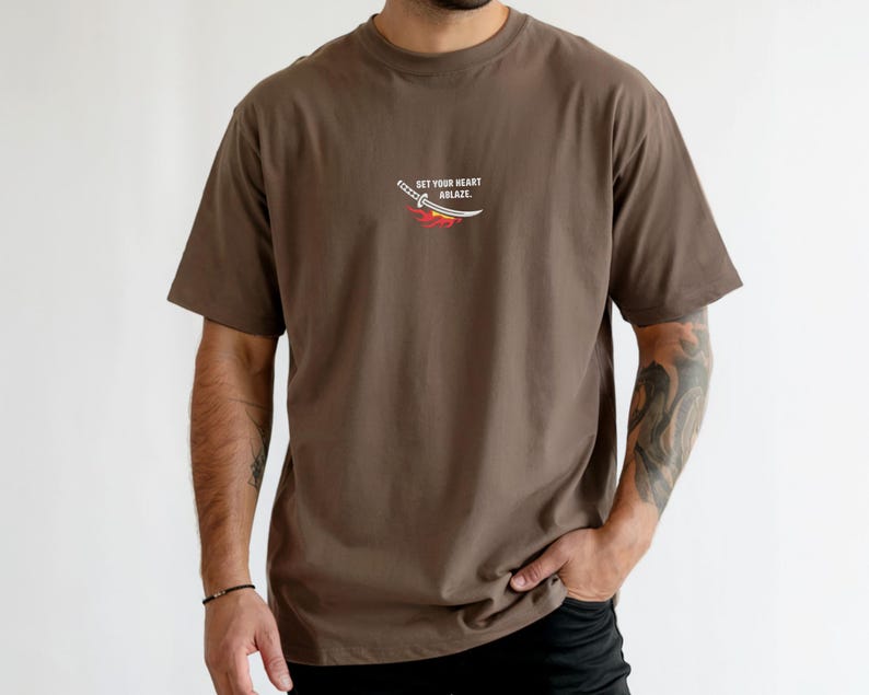 Puede incluir: Una camiseta marr&oacute;n de manga corta con un peque&ntilde;o gr&aacute;fico en el pecho. El gr&aacute;fico muestra una espada katana blanca sobre una llama estilizada roja y naranja, con el texto "SET YOUR HEART ABLAZE" impreso en letras may&uacute;sculas blancas.