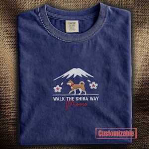 以下が含まれることがあります： ネイビーブルーのTシャツ。茶色の柴犬、白い山の頂、桜の花が刺繍されています。テキストは「WALK THE SHIBA WAY」と赤字の筆記体で「Momo」と書かれています。襟の内側のラベルには「COMFORT COLORS」と記されています。