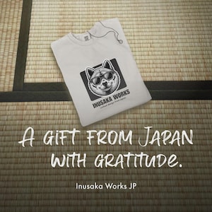 May include: White t-shirt with a graphic of a dog wearing sunglasses, framed in a black square. The text "INUSAKA WORKS" and "crafted design from Japan" are below the dog graphic. The text "A GIFT FROM JAPAN WITH GRATITUDE" is below the t-shirt.