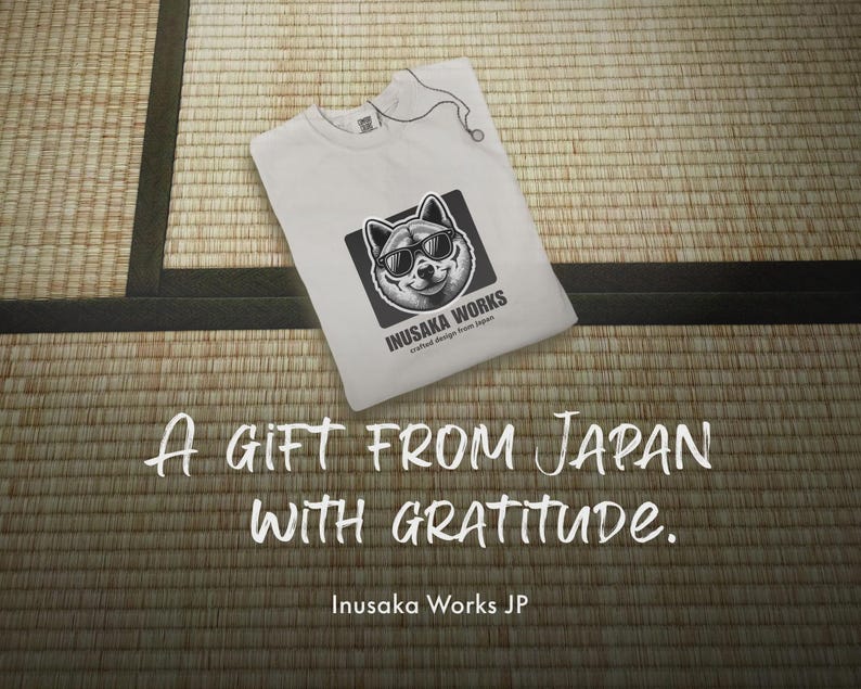 May include: White t-shirt with a graphic of a dog wearing sunglasses, framed in a black square. The text "INUSAKA WORKS" and "crafted design from Japan" are below the dog graphic. The text "A GIFT FROM JAPAN WITH GRATITUDE" is below the t-shirt.