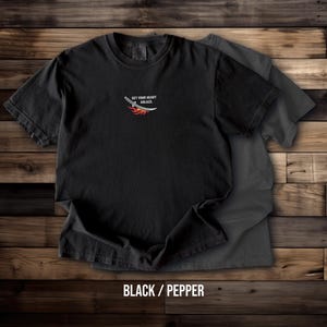 Puede incluir: Una camiseta negra de manga corta con un peque&ntilde;o gr&aacute;fico en el pecho de una espada con llamas y el texto "SET YOUR HEART ABLAZY." Una segunda camiseta de color gris carb&oacute;n es parcialmente visible debajo de la camiseta negra principal.