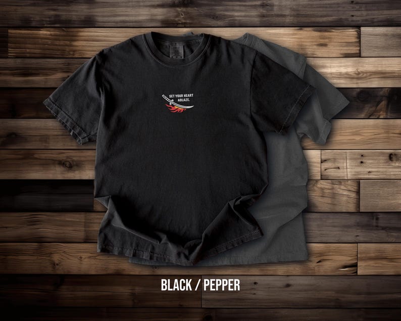Puede incluir: Una camiseta negra de manga corta con un peque&ntilde;o gr&aacute;fico en el pecho de una espada con llamas y el texto "SET YOUR HEART ABLAZY." Una segunda camiseta de color gris carb&oacute;n es parcialmente visible debajo de la camiseta negra principal.