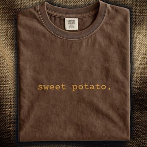 以下が含まれることがあります： 「sweet potato」の刺繍が入った、折りたたまれた茶色のTシャツ。クルーネックで、襟元には小さなタグが付いています。生地は柔らかく、着心地が良さそうです。
