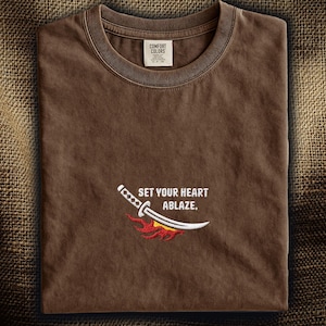Puede incluir: Una camiseta marr&oacute;n de cuello redondo con un dise&ntilde;o bordado de una espada katana blanca con un gr&aacute;fico de llamas rojas y naranjas debajo. El texto "SET YOUR HEART ABLAZE." est&aacute; bordado en blanco sobre la espada. Se ve una etiqueta Comfort Colors.