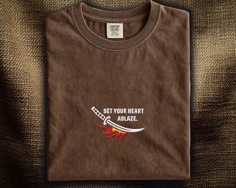 Puede incluir: Una camiseta marr&oacute;n de cuello redondo con un dise&ntilde;o bordado de una espada katana blanca con un gr&aacute;fico de llamas rojas y naranjas debajo. El texto "SET YOUR HEART ABLAZE." est&aacute; bordado en blanco sobre la espada. Se ve una etiqueta Comfort Colors.