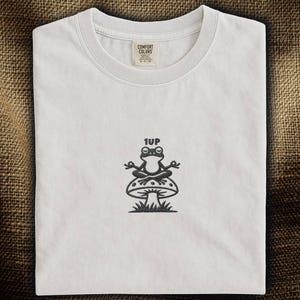 May include: White t-shirt with a crew neck and a graphic of a meditating frog on a mushroom, with the text "1UP" above. The shirt has a tag that reads "Comfort Colors".