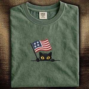 以下が含まれることがあります： 小さなアメリカ国旗を持った黒猫の刺繍デザインが施された緑色のTシャツ。Tシャツはクルーネックで、「Comfort Colors」と書かれたタグが付いています。
