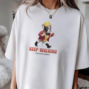 May include: Cream-colored t-shirt featuring a cartoon French Bulldog in a red jacket, yellow hat, and black shoes, holding a cane. The text "KEEP WALKING - Frenchie Walker -" is printed below the image.