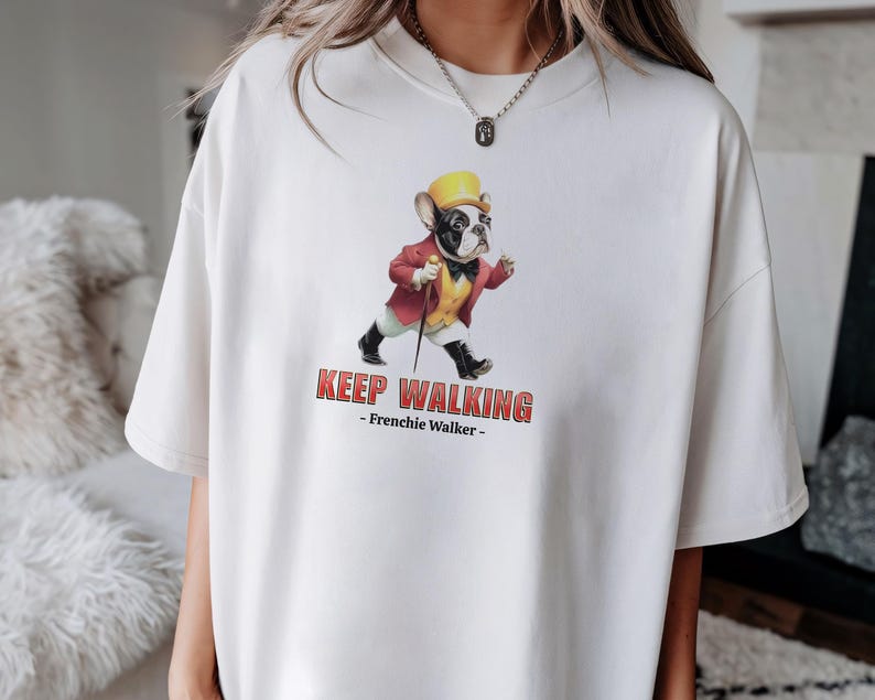 May include: Cream-colored t-shirt featuring a cartoon French Bulldog in a red jacket, yellow hat, and black shoes, holding a cane. The text "KEEP WALKING - Frenchie Walker -" is printed below the image.