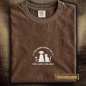 以下が含まれることがあります： 「YOU MAKE EVERY DAY FEEL LIKE A HOLIDAY」の文字と犬と猫のイラストが描かれた茶色のTシャツ。クルーネックで、「COMFORT COLORS」のタグが付いています。下部には「Customizable」の文字があります。