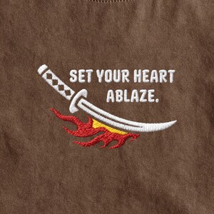 Puede incluir: Una superficie de tela marr&oacute;n con un dise&ntilde;o bordado de una espada katana blanca situada sobre llamas estilizadas rojas y amarillas, acompa&ntilde;ada por el texto bordado en blanco que dice, SET YOUR HEART ABLAZE.