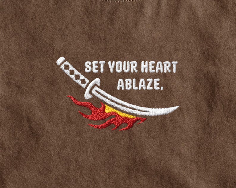 Puede incluir: Una superficie de tela marr&oacute;n con un dise&ntilde;o bordado de una espada katana blanca situada sobre llamas estilizadas rojas y amarillas, acompa&ntilde;ada por el texto bordado en blanco que dice, SET YOUR HEART ABLAZE.