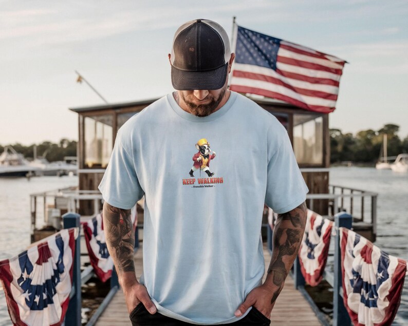 May include: Light blue t-shirt featuring a cartoon bulldog wearing a yellow hat and red jacket, walking with a cane. The text "Keep Walking" is below the graphic. A black and gray trucker hat is worn.