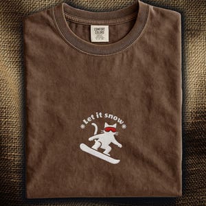 以下が含まれることがあります： サングラスをかけた猫がスノーボードをしている白いグラフィックの茶色のTシャツ。「Let it snow」の文字が猫の上にあります。シャツはクルーネックで、上部中央にタグが付いています。