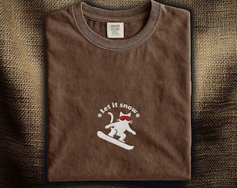 Könnte beinhalten: Ein braunes Kurzarm-T-Shirt mit einer Stickerei, die eine weiße Katze mit roter Skibrille auf einem Snowboard zeigt. Der Text "Let it snow" ist bogenförmig über der Katze angebracht und von zwei kleinen Schneeflocken flankiert. Rundhalsausschnitt.