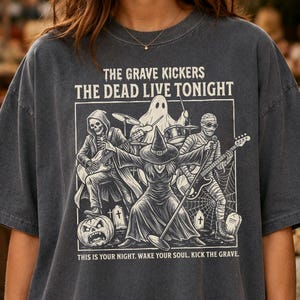 May include: A charcoal gray t-shirt featuring a graphic print of a band of monsters playing instruments. The text reads THE GRAVE KICKERS THE DEAD LIVE TONIGHT and THIS IS YOUR NIGHT. WAKE YOUR SOUL. KICK THE GRAVE. The design includes a skeleton, ghost, mummy, and witch.