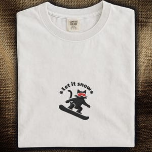 以下が含まれることがあります： 赤いゴーグルをつけた黒猫がスノーボードをしている白いTシャツ。「Let it snow」の文字が猫の上にあります。シャツにはComfort Colorsのタグが付いています。