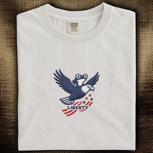 以下が含まれることがあります： 白いTシャツ。ネイビーと白の刺繍デザインで、カエルの目のワシが翼を広げ、「LIBERTY」と赤と白で書かれたバナーを持っています。シャツは茶色の表面に折りたたまれています。