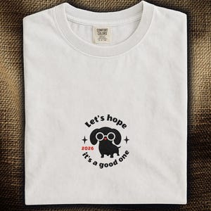 以下が含まれることがあります： クルーネックの白いTシャツ。Comfort Colorsのタグが付いています。メガネをかけた黒いダックスフンドが描かれており、「Let's hope it's a good one」というテキストと「2026」の年号が赤色で表示されています。