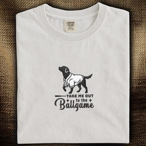 以下が含まれることがあります： 野球のユニフォームを着た犬の黒いグラフィックが描かれた白いTシャツ。「TAKE ME OUT to the Ballgame」の文字が下にあります。シャツにはComfort Colorsのタグが付いています。