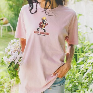May include: A light pink t-shirt featuring a cartoon illustration of a French Bulldog wearing a hat and coat, walking with a cane. The text "KEEP WALKING - Frenchie Walker" is printed below the illustration. The shirt is made of soft material.