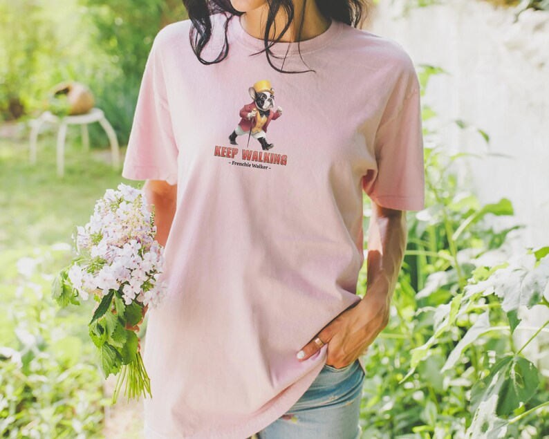 May include: A light pink t-shirt featuring a cartoon illustration of a French Bulldog wearing a hat and coat, walking with a cane. The text "KEEP WALKING - Frenchie Walker" is printed below the illustration. The shirt is made of soft material.