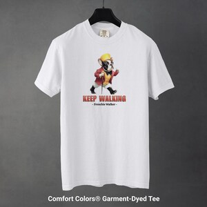 May include: White Comfort Colors garment-dyed tee featuring a cartoon French Bulldog dressed in a red jacket, yellow hat, and black shoes, walking with a cane. The text "KEEP WALKING - Frenchie Walker-" is printed in red.