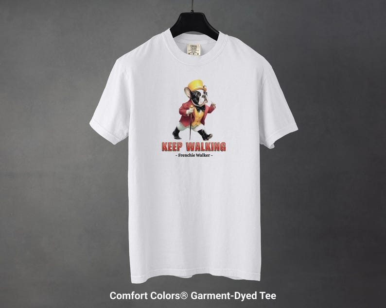 May include: White Comfort Colors garment-dyed tee featuring a cartoon French Bulldog dressed in a red jacket, yellow hat, and black shoes, walking with a cane. The text "KEEP WALKING - Frenchie Walker-" is printed in red.