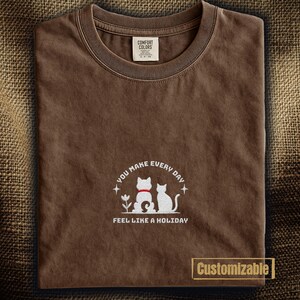 以下が含まれることがあります： 丸首の茶色のTシャツ。2匹の猫の白いグラフィックと「YOU MAKE EVERY DAY FEEL LIKE A HOLIDAY」というテキストが特徴です。「Comfort Colors」のタグが付いています。「Customizable」という単語も表示されています。