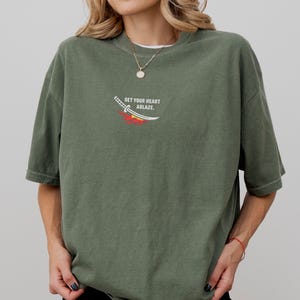 Puede incluir: Una camiseta de color verde oliva con un peque&ntilde;o gr&aacute;fico en el pecho. El gr&aacute;fico muestra una espada blanca con un dise&ntilde;o de llamas rojas y amarillas debajo, acompa&ntilde;ado del texto "SET YOUR HEART ABLAZE." en letras may&uacute;sculas blancas.