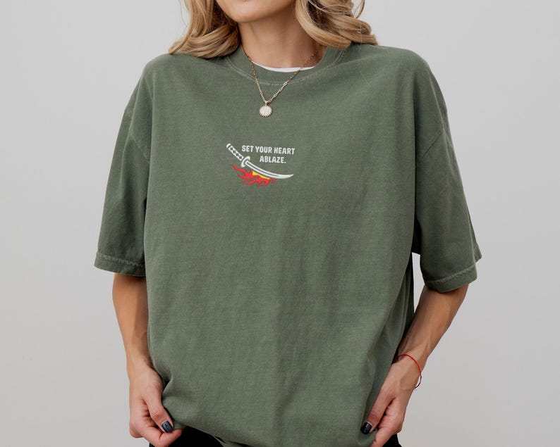 Puede incluir: Una camiseta de color verde oliva con un peque&ntilde;o gr&aacute;fico en el pecho. El gr&aacute;fico muestra una espada blanca con un dise&ntilde;o de llamas rojas y amarillas debajo, acompa&ntilde;ado del texto "SET YOUR HEART ABLAZE." en letras may&uacute;sculas blancas.