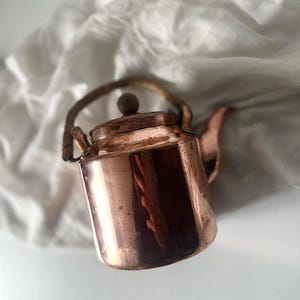 May include: A copper tea kettle with a wooden handle and a small wooden knob on the lid. The kettle has a spout and is set against a white background.