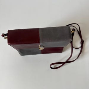 May include: A rectangular shoulder bag with a color block design. The bag features a burgundy and gray color scheme, with a gold-tone clasp and a matching burgundy strap. The bag is made of leather.