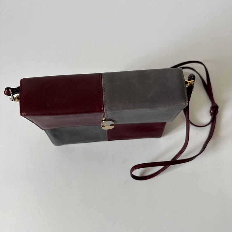 May include: A rectangular shoulder bag with a color block design. The bag features a burgundy and gray color scheme, with a gold-tone clasp and a matching burgundy strap. The bag is made of leather.