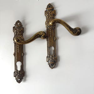 Può includere: Due antiche maniglie per porte in ottone con dettagli floreali ornati. Le maniglie hanno un design curvo e ritagli per la serratura. L'ottone ha una calda patina invecchiata, che suggerisce l'artigianato vintage.
